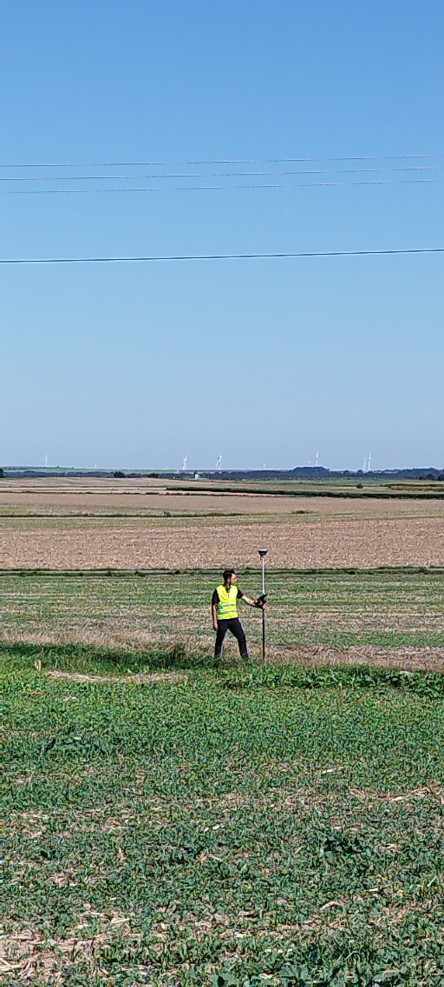 Geodeta w żółtej kamizelce mierzy pole za pomocą precyzyjnego instrumentu geodezyjnego, w tle widać turbiny wiatrowe i błękitne niebo.