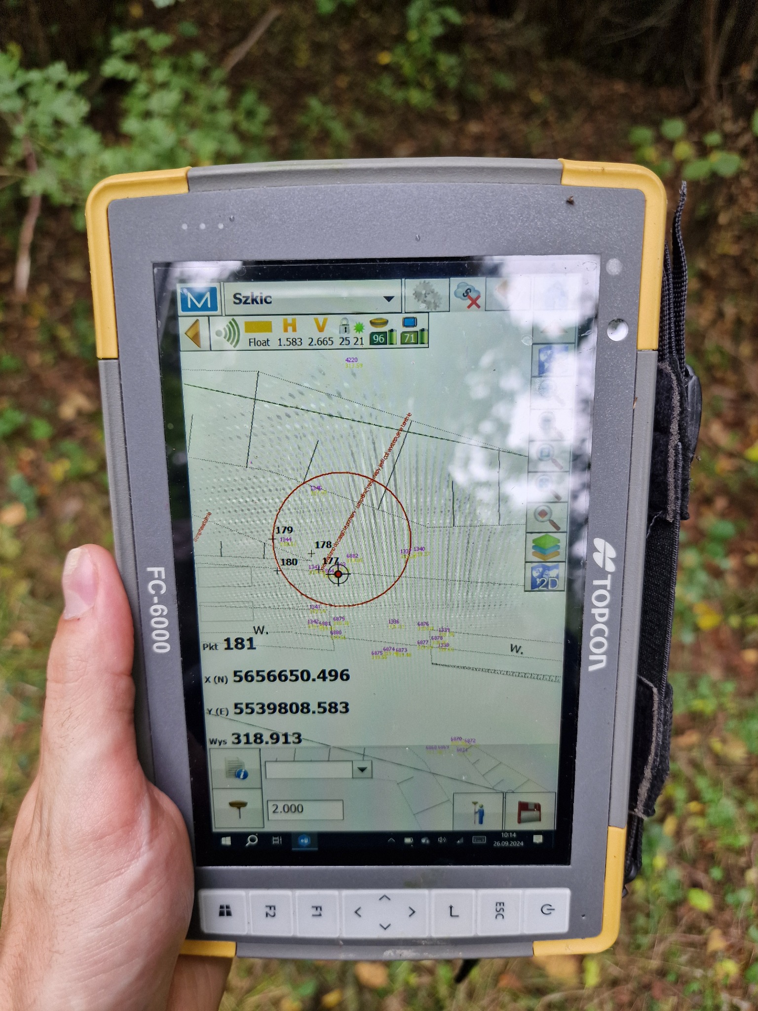 Ekran tabletu Topcon FC-6000 wyświetlający mapę z zaznaczonymi punktami pomiarowymi w terenie, trzymany w dłoni na tle zieleni.