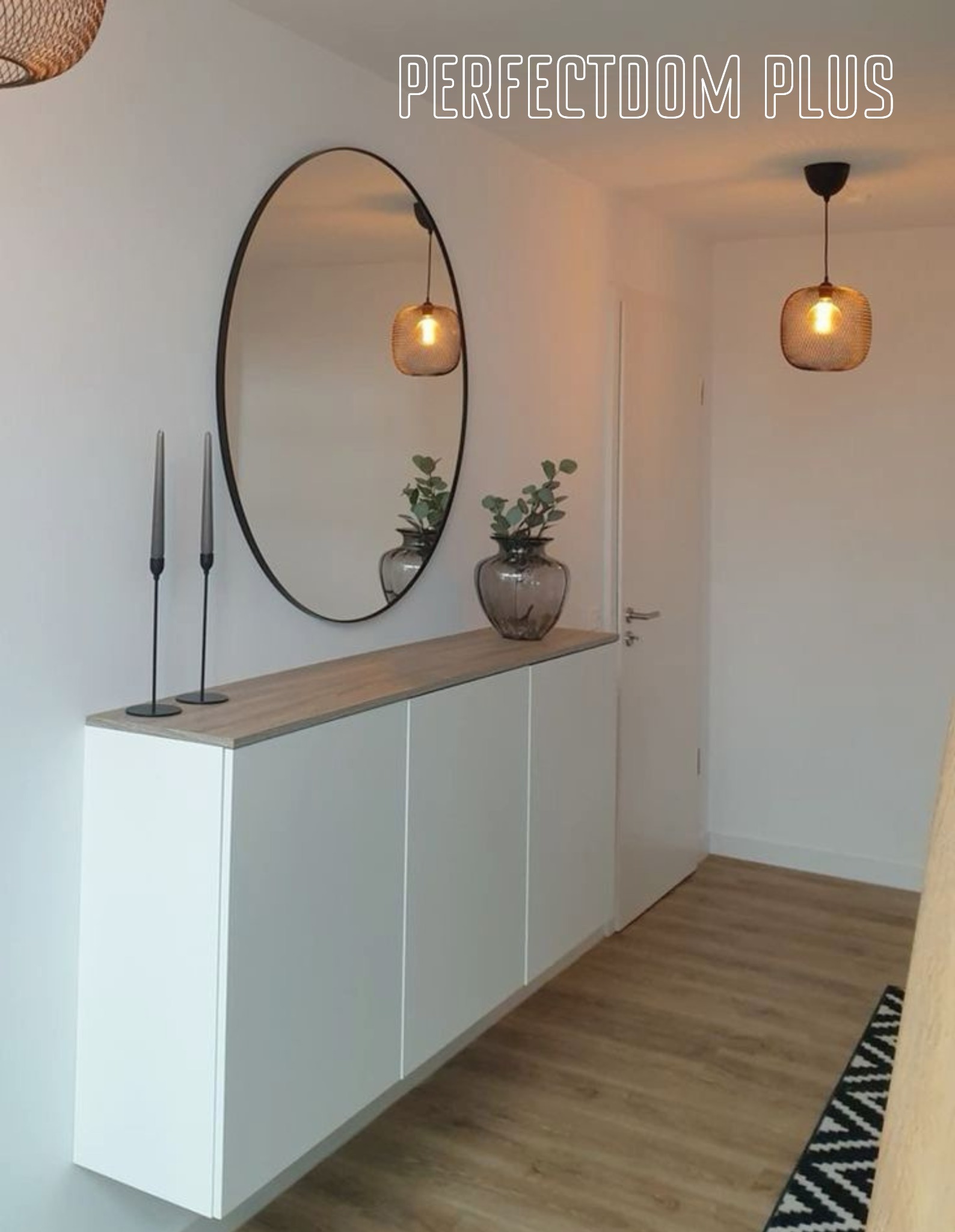 Nowoczesny przedpokój z białymi szafkami wiszącymi, blatem w kolorze jasnego drewna, okrągłym lustrem i szarymi świecami, podkreślony wiszącymi lampami w stylu industrialnym.