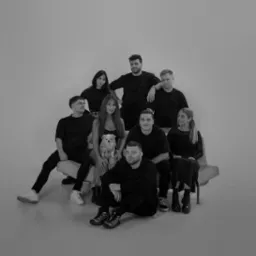 Czarno-białe zdjęcie grupowe dziewięciu osób i małego psa rasy szpic miniaturowy, siedzących na jasnej sofie i krzesłach na jednolitym tle.