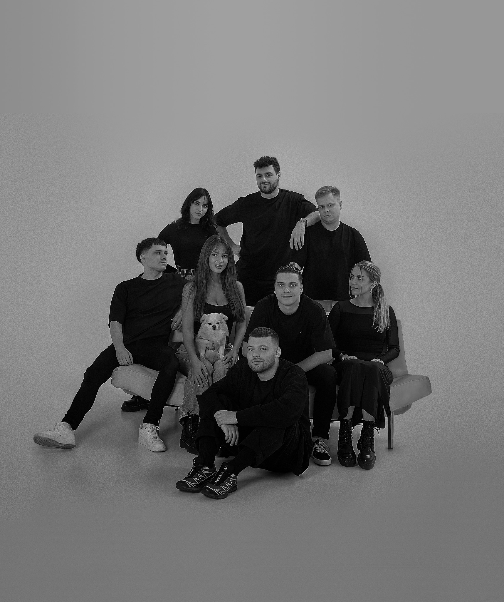 Czarno-białe zdjęcie grupowe dziewięciu osób i małego psa rasy szpic miniaturowy, siedzących na jasnej sofie i krzesłach na jednolitym tle.