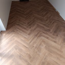 Usługi wykończeniowe - Ułożony parkiet w jodełkę na podłodze, widoczne paczki paneli przy ścianie, fragment surowej ściany.