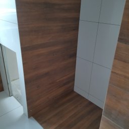 Usługi wykończeniowe - Wykończona łazienka z brązowymi płytkami imitującymi drewno na podłodze i części ściany oraz białymi, kwadratowymi płytkami na pozostałej części ściany.