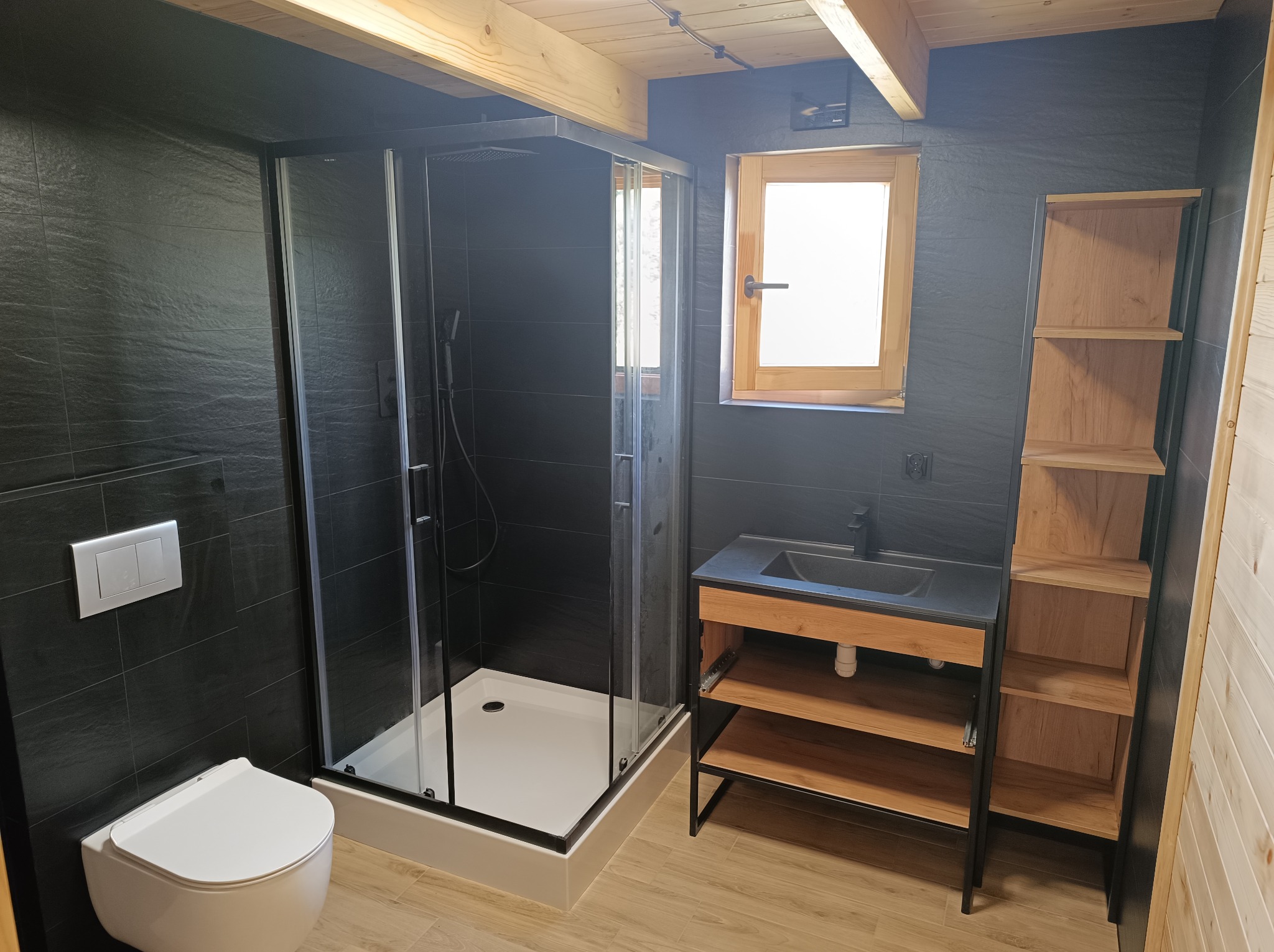 Nowoczesna łazienka z czarnymi płytkami imitującymi kamień, kabiną prysznicową typu walk-in z czarnymi profilami, podwieszaną toaletą, drewnianą szafką z czarną umywalką i drewnianym regałem...