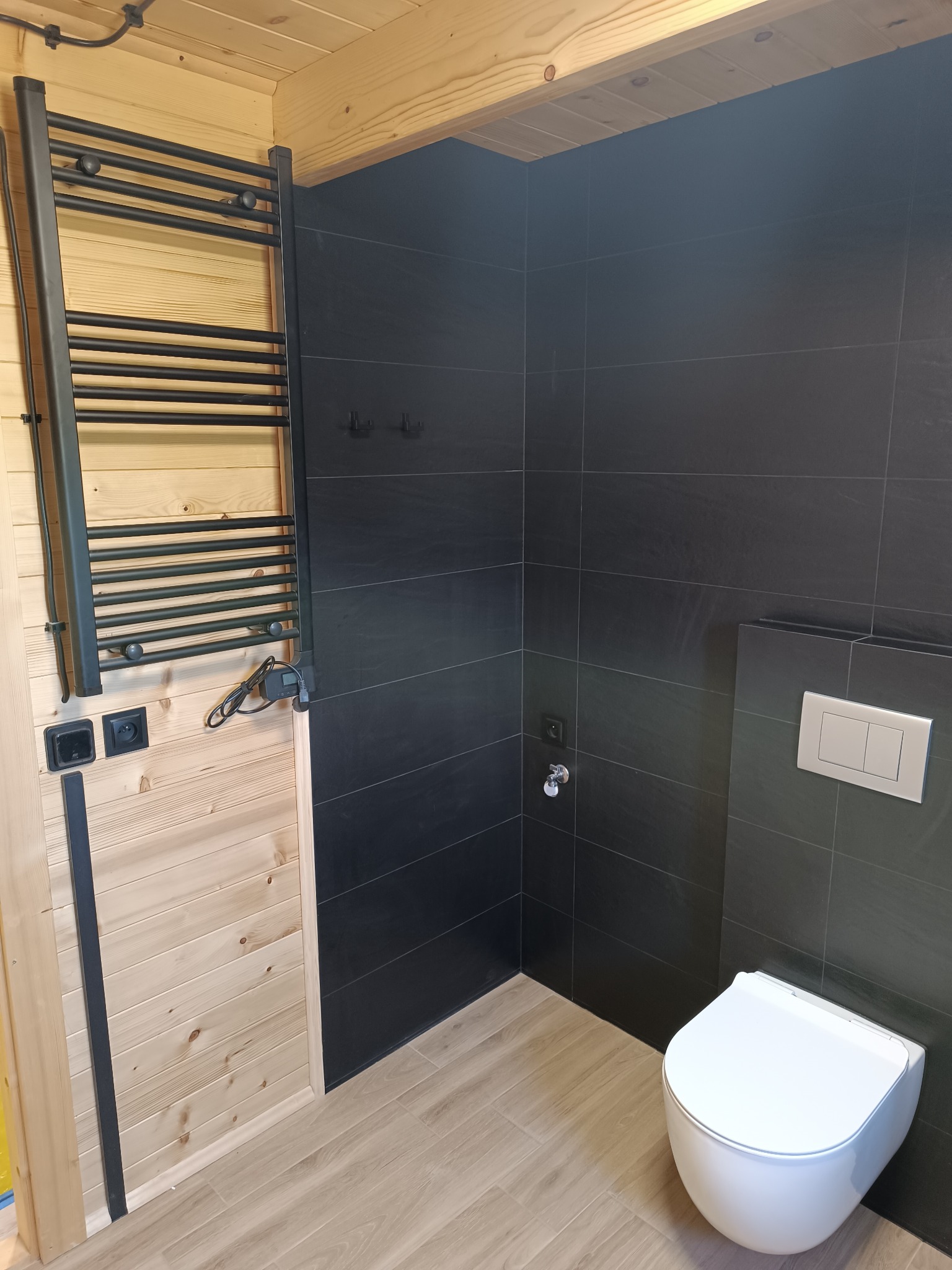 Nowoczesna łazienka z czarnymi płytkami, drewnianą boazerią i podwieszaną toaletą, grzejnik drabinkowy w kolorze czarnym.