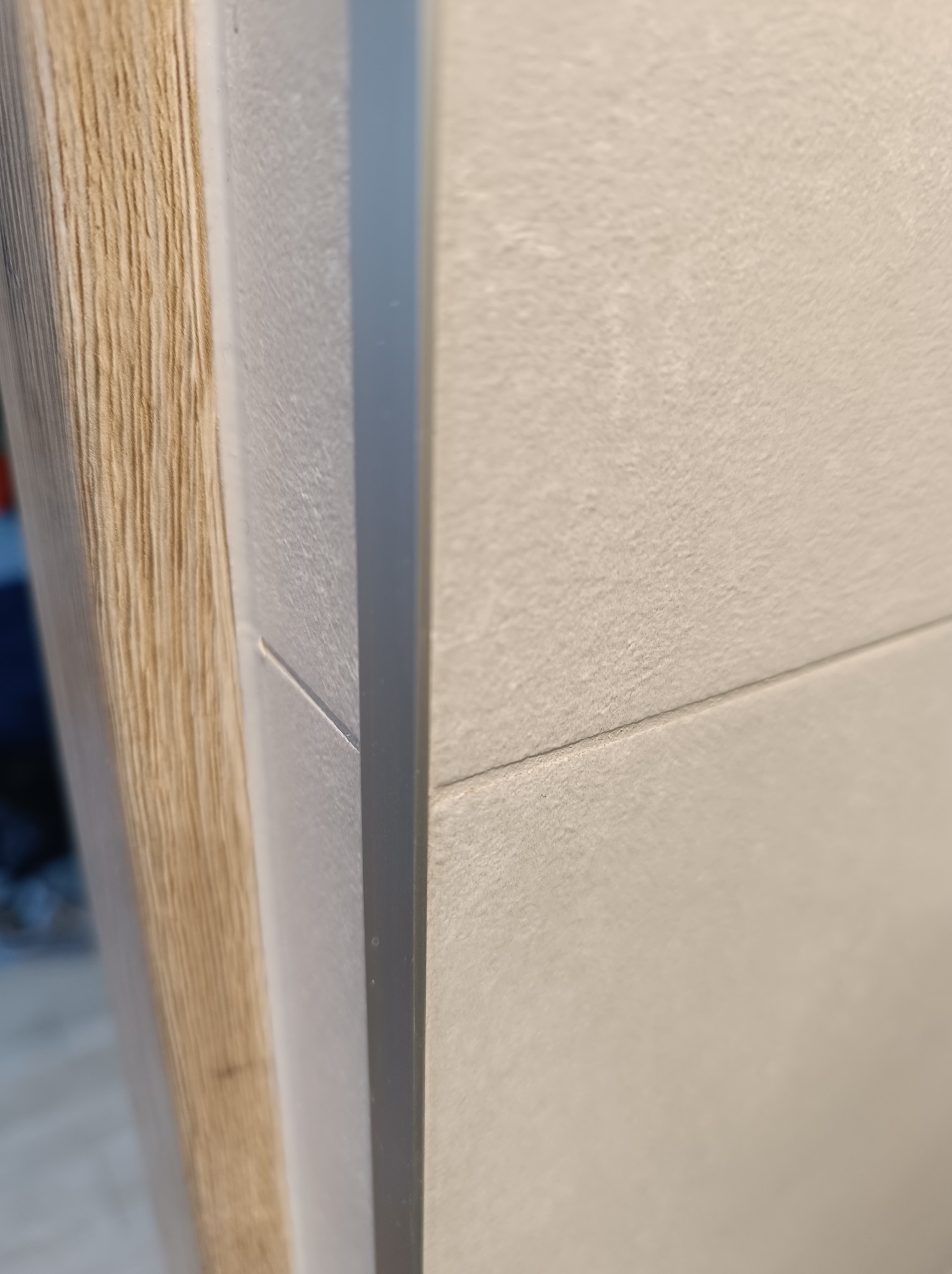 Detal wykończenia ściany z jasnych, matowych płytek ceramicznych, połączonych szarą fugą, obok drewnopodobnej ramy drzwi wykończonej szarą listwą.