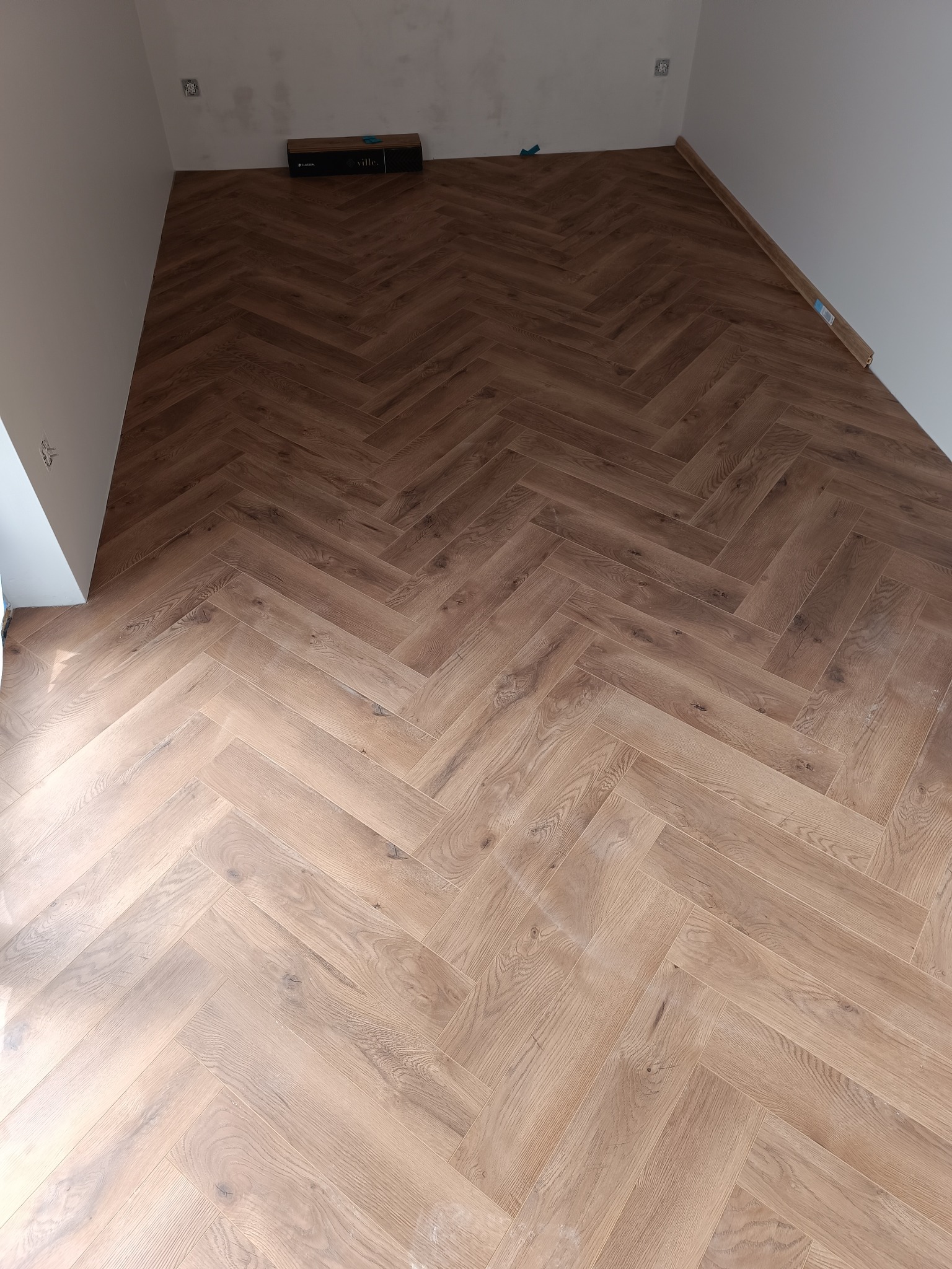 Ułożony parkiet w jodełkę na podłodze, widoczne paczki paneli przy ścianie, fragment surowej ściany.