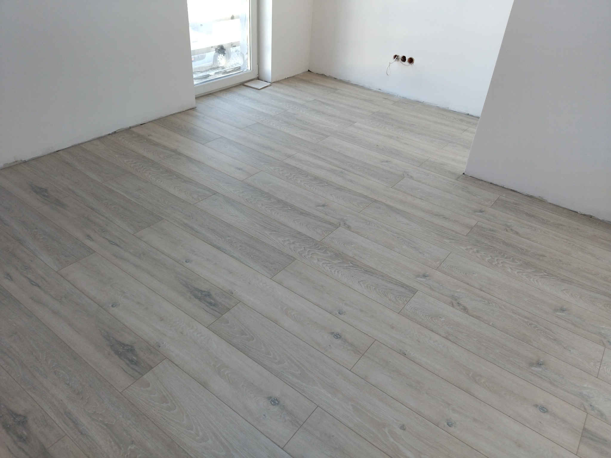Jasnoszara podłoga z paneli ułożona diagonalnie w pustym, odnowionym pokoju z białymi ścianami i gniazdkami elektrycznymi bez osprzętu.