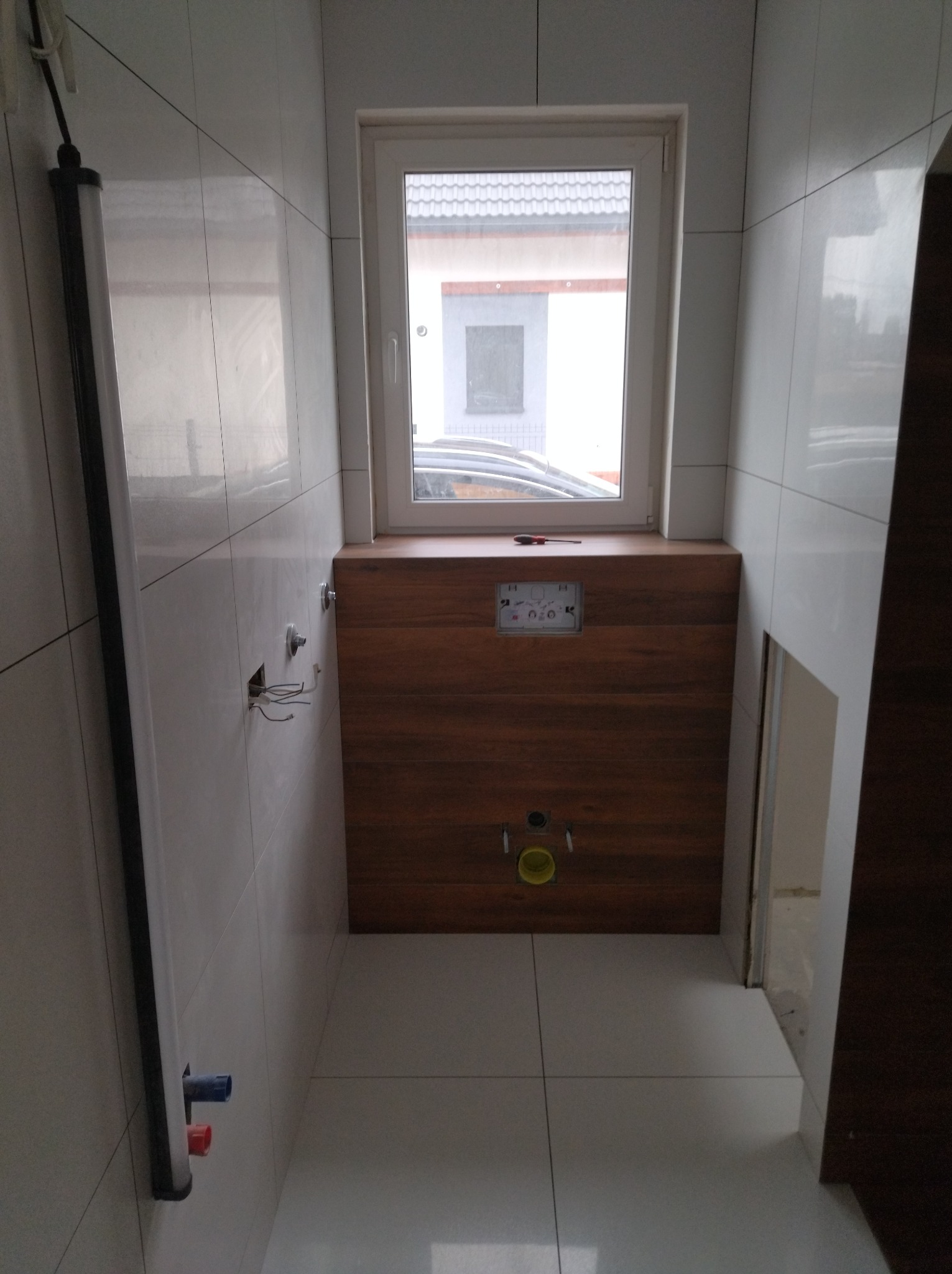 Wnętrze łazienki w trakcie remontu: białe płytki na ścianach i podłodze, zabudowa WC z drewnopodobną okładziną, widoczne instalacje wodno-kanalizacyjne, okno z widokiem na dom.