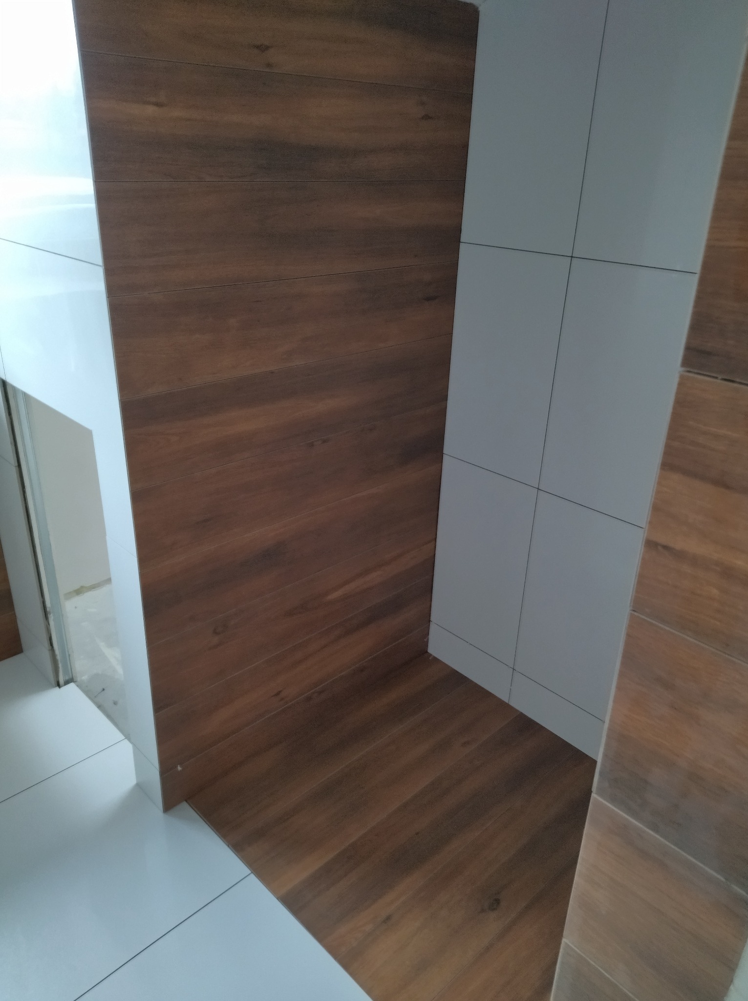 Wykończona łazienka z brązowymi płytkami imitującymi drewno na podłodze i części ściany oraz białymi, kwadratowymi płytkami na pozostałej części ściany.