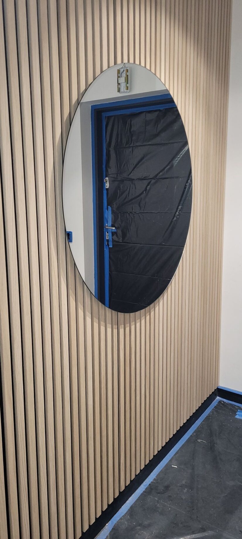 Ściana z drewnianych lameli z owalnym lustrem, widoczne zabezpieczenie folią i taśmą malarską, prawdopodobnie w trakcie remontu.