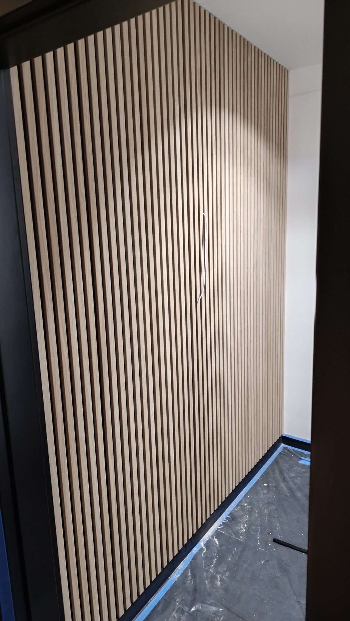 Ściana wykończona panelami lamelowymi z widocznym kablem wystającym ze ściany, zabezpieczona folią malarską na podłodze.