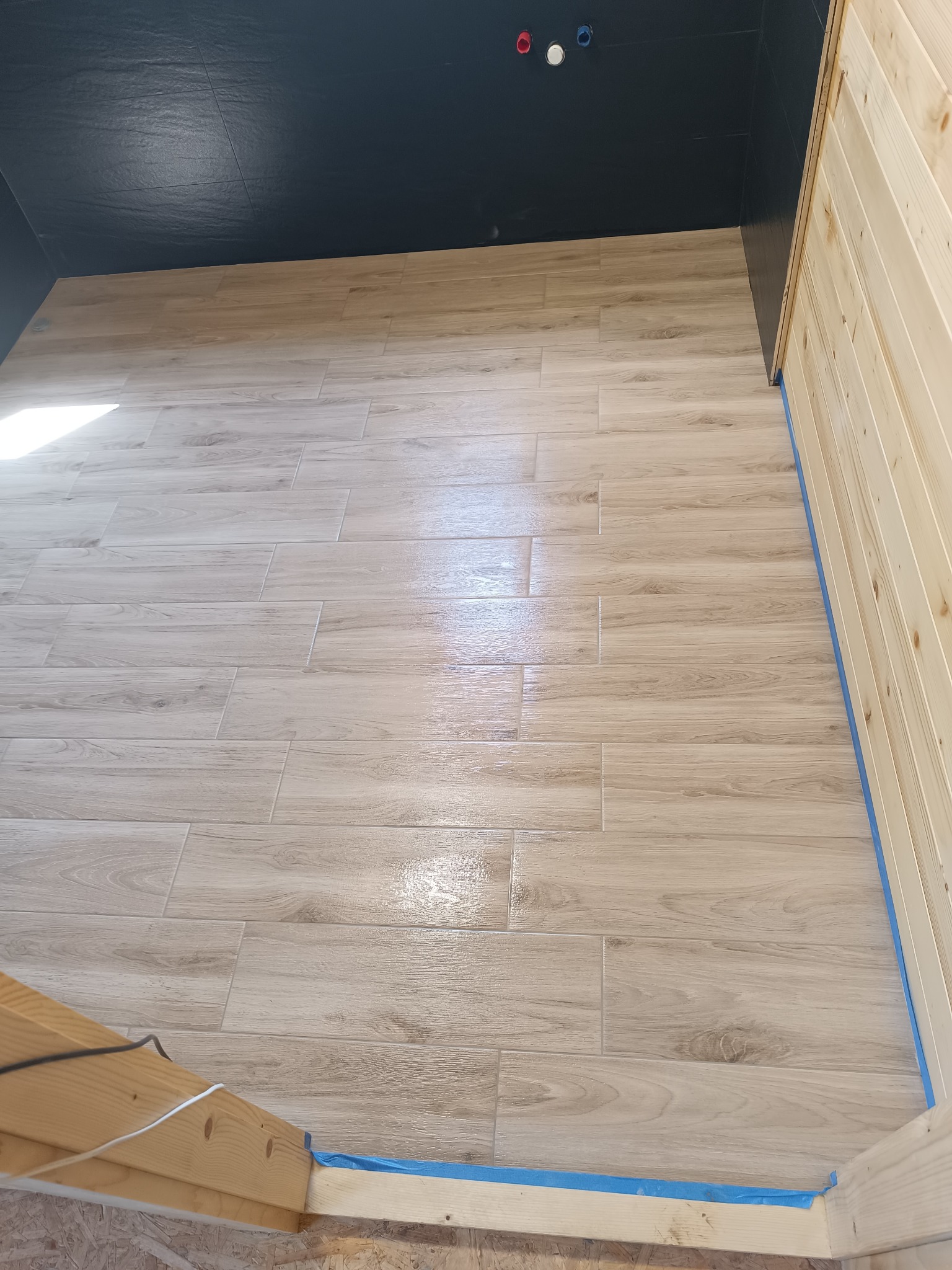 Ułożona podłoga z płytek imitujących drewno w jasnym odcieniu, ściany wykończone ciemnymi płytkami i boazerią, zabezpieczone niebieską taśmą malarską.