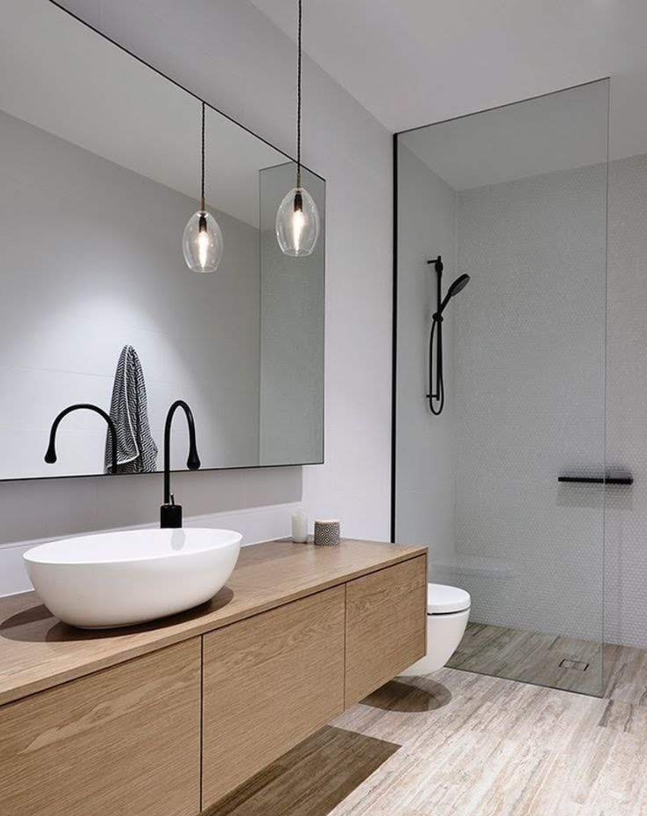 Nowoczesna łazienka z drewnianą szafką pod umywalką, owalną białą umywalką nablatową, czarną armaturą i szklaną kabiną prysznicową.