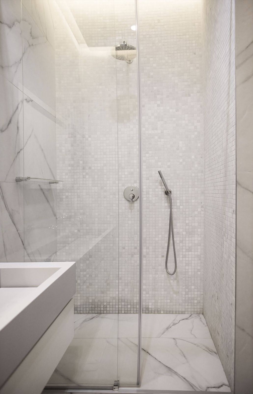 Nowoczesna kabina prysznicowa z deszczownicą i słuchawką, wykończona białymi mozaikami i płytami imitującymi marmur, z minimalistyczną umywalką po lewej stronie.