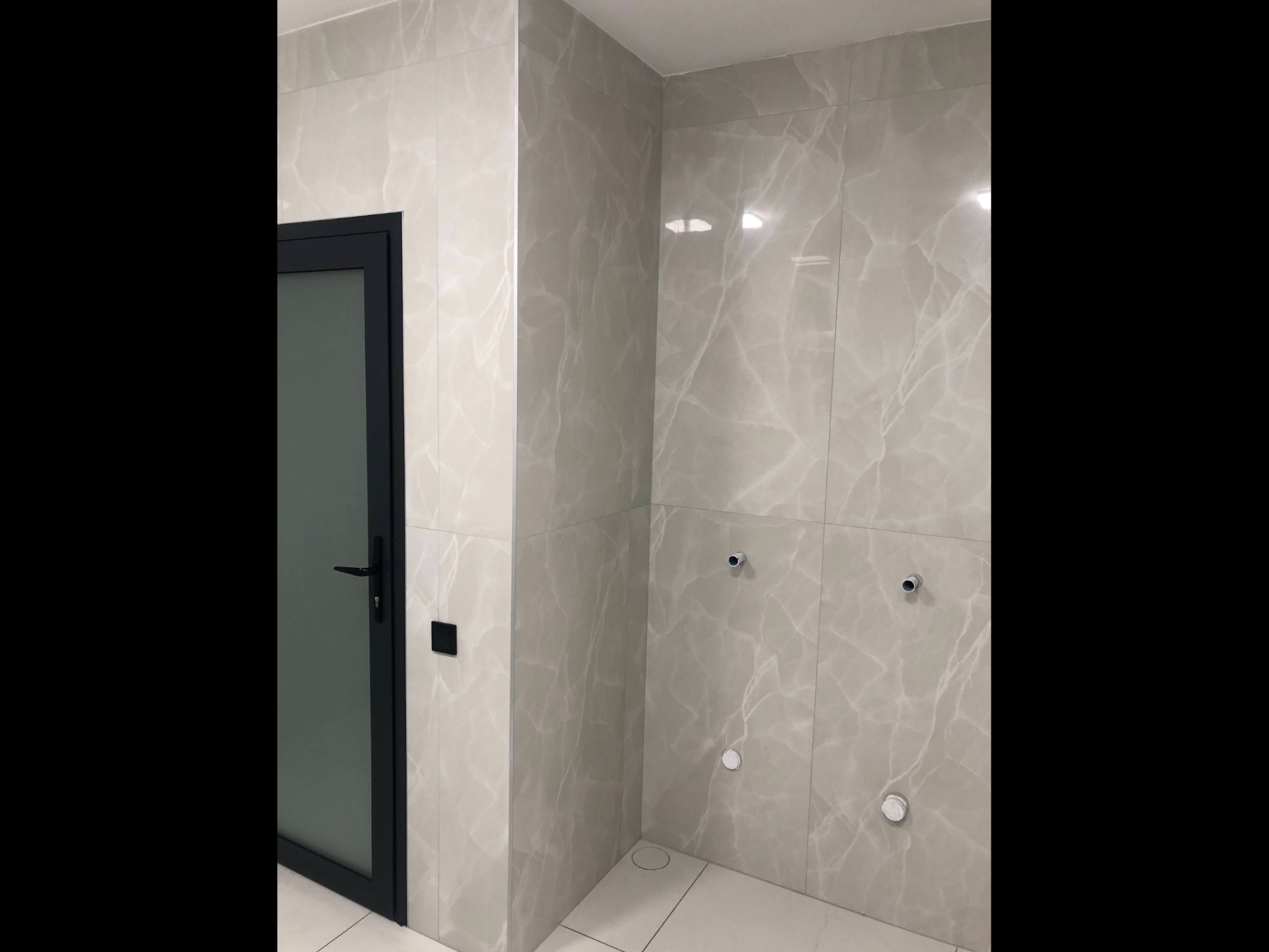 Wykończona ściana łazienki z płytkami imitującymi marmur, widoczne podejścia wodno-kanalizacyjne i czarne drzwi z klamką.