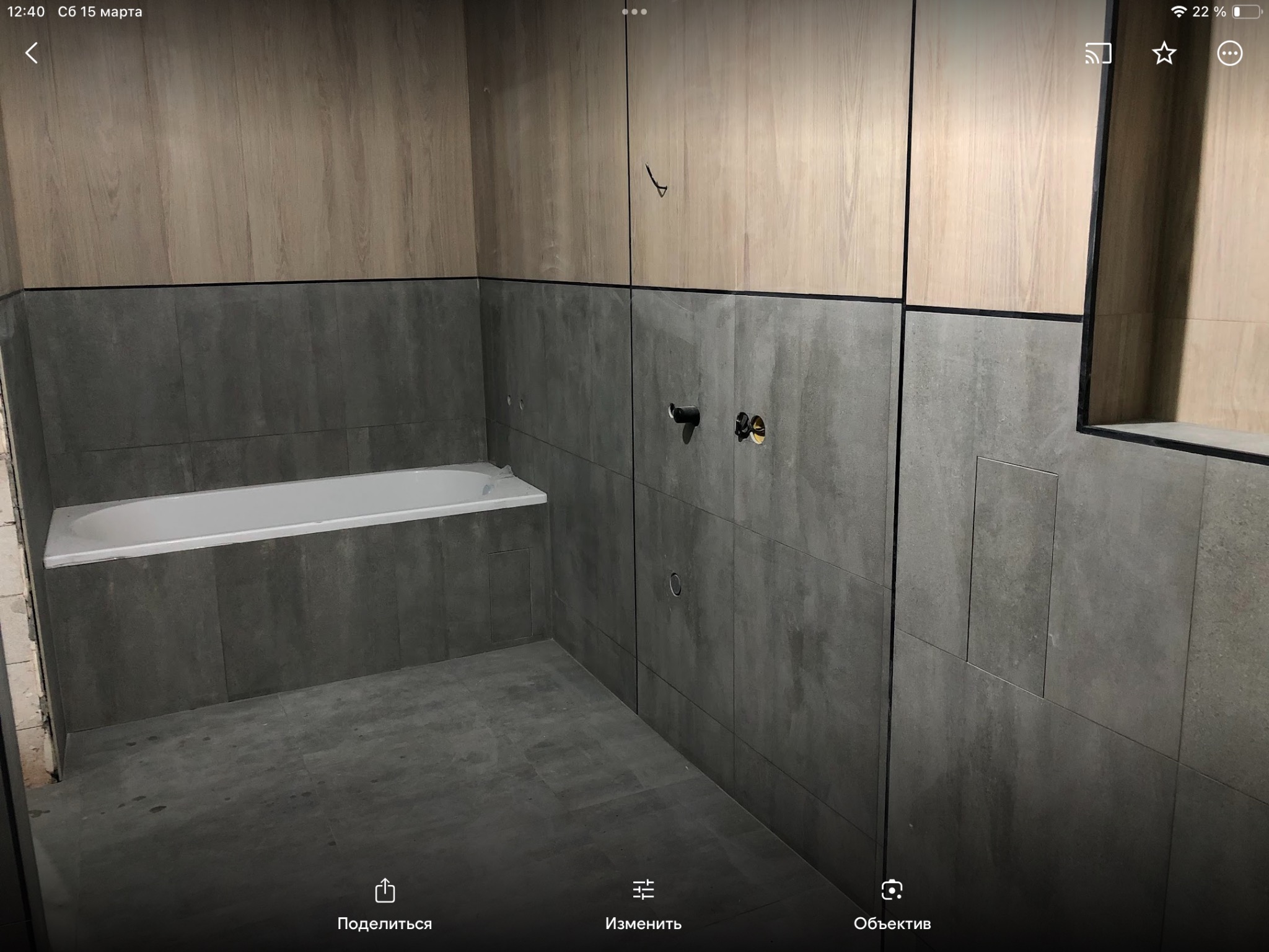 Wnętrze łazienki w trakcie wykańczania: szare płytki na ścianach i podłodze, wanna w rogu, drewnopodobne panele na górnej części ścian, widoczne instalacje wodne.