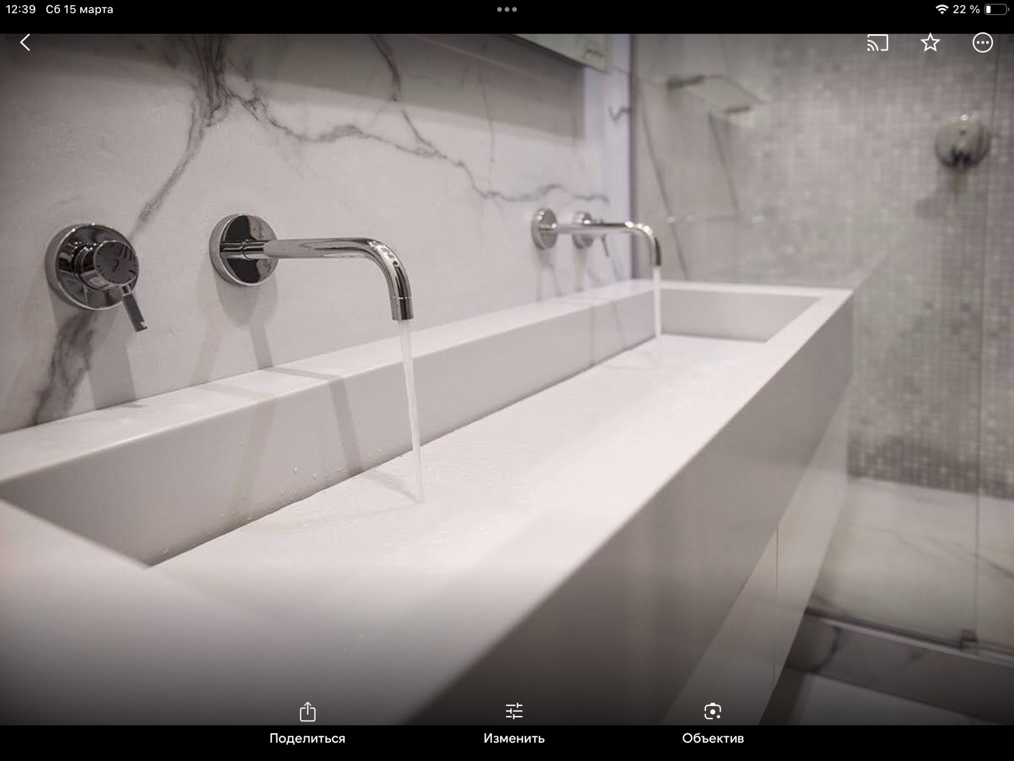 Nowoczesna łazienka z długą, białą, prostokątną umywalką wpuszczoną w blat, chromowane baterie ścienne, marmurowe płyty na ścianie i mozaika pod prysznicem.