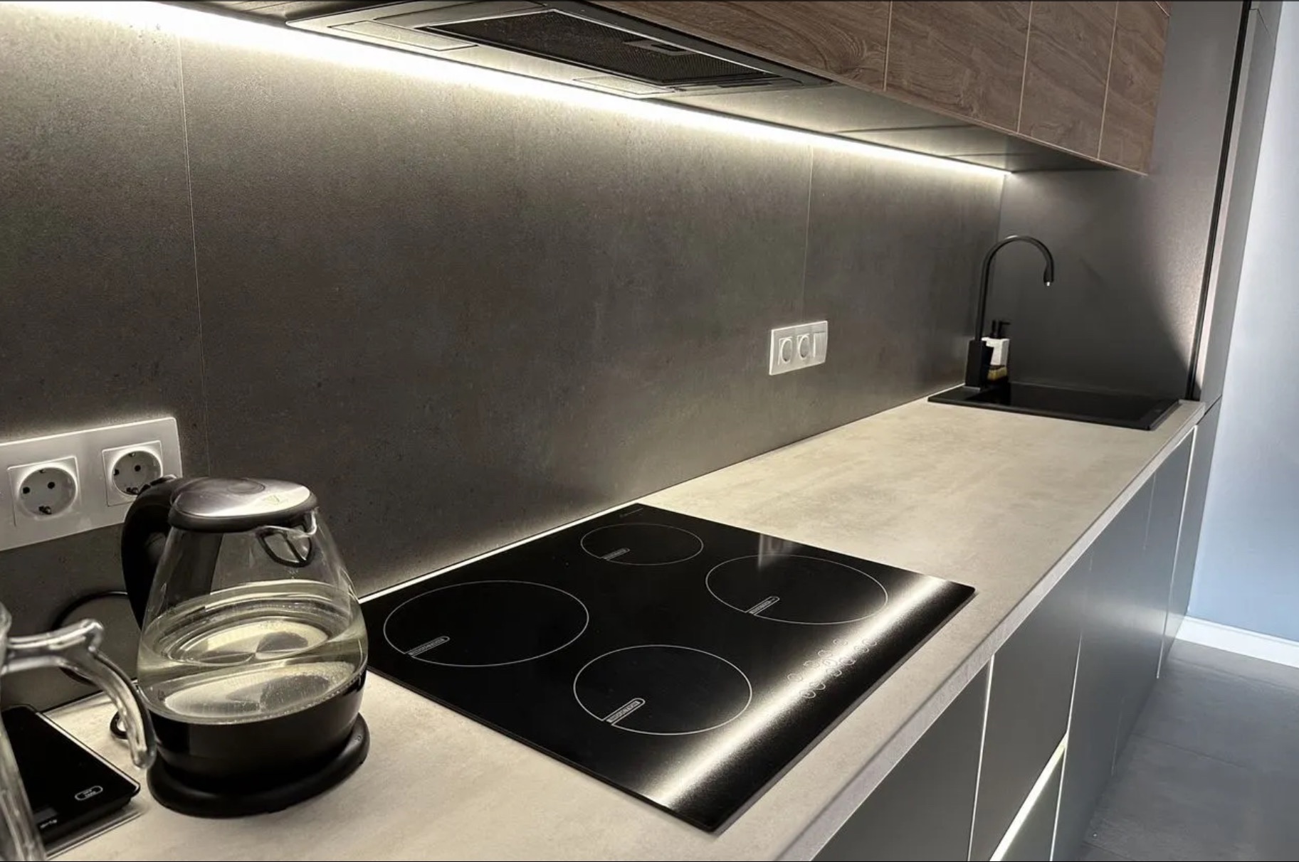 Nowoczesna kuchnia z indukcyjną płytą grzewczą i minimalistycznym oświetleniem LED pod szafkami. Eleganckie wykończenie ścian i blatów w odcieniach szarości.