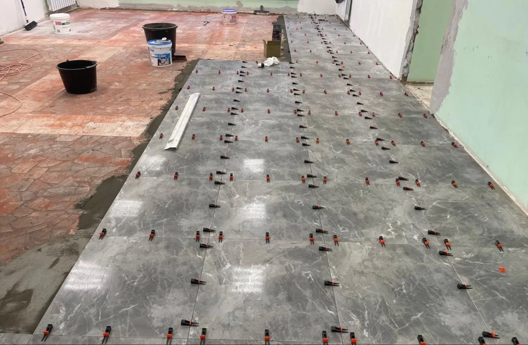 Układanie szarych płytek na podłodze z użyciem klipsów poziomujących. Fragment starej, czerwonej podłogi widoczny obok nowej posadzki. Prace w toku.