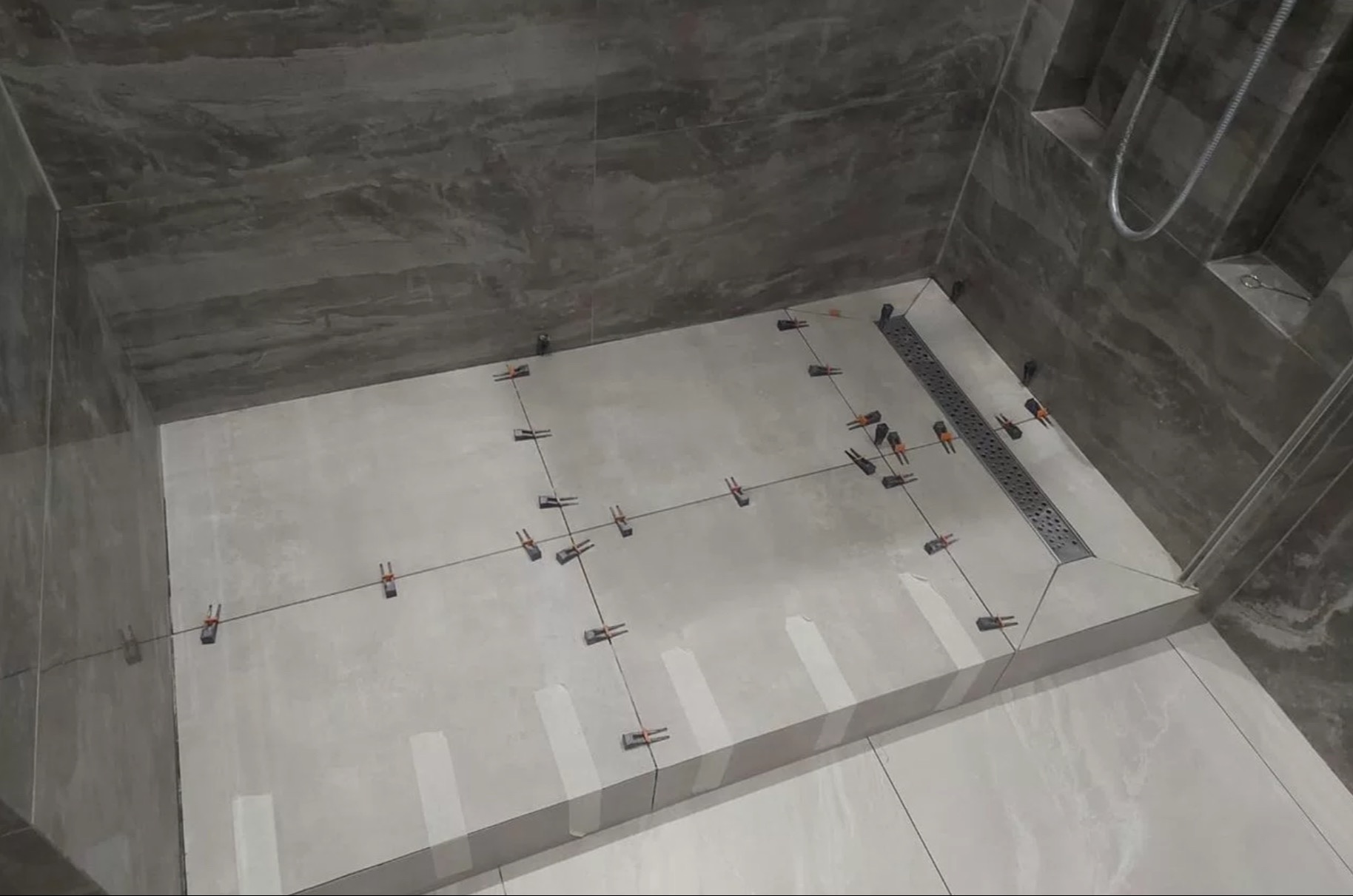 Białe płytki podłogowe w kabinie prysznicowej, wyrównane klipsami i sznurkiem. Widoczny odpływ liniowy i marmurowe płytki ścienne, częściowo ułożone.