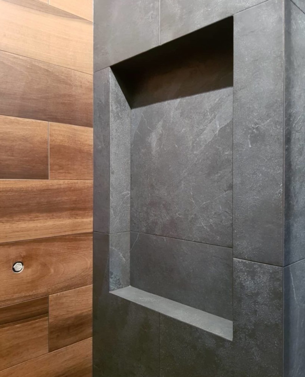 Wnęka wyłożona szarą płytką w kabinie prysznicowej. Na ścianie obok widoczna okładzina imitująca drewno. Nowoczesne wykończenie.