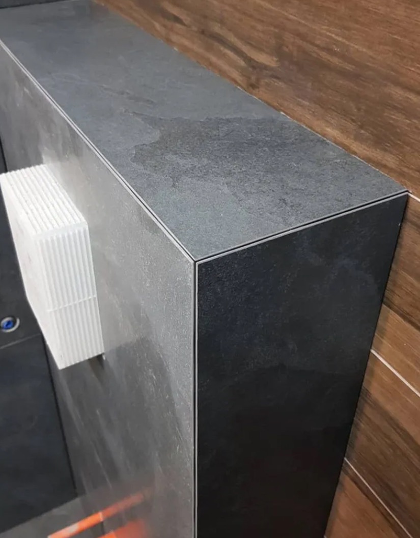 Wykończony fragment ściany z ciemnych, matowych płytek, z widocznym narożnikiem i elementem wentylacyjnym. Ujęcie z bliska, podkreślające precyzję wykonania.