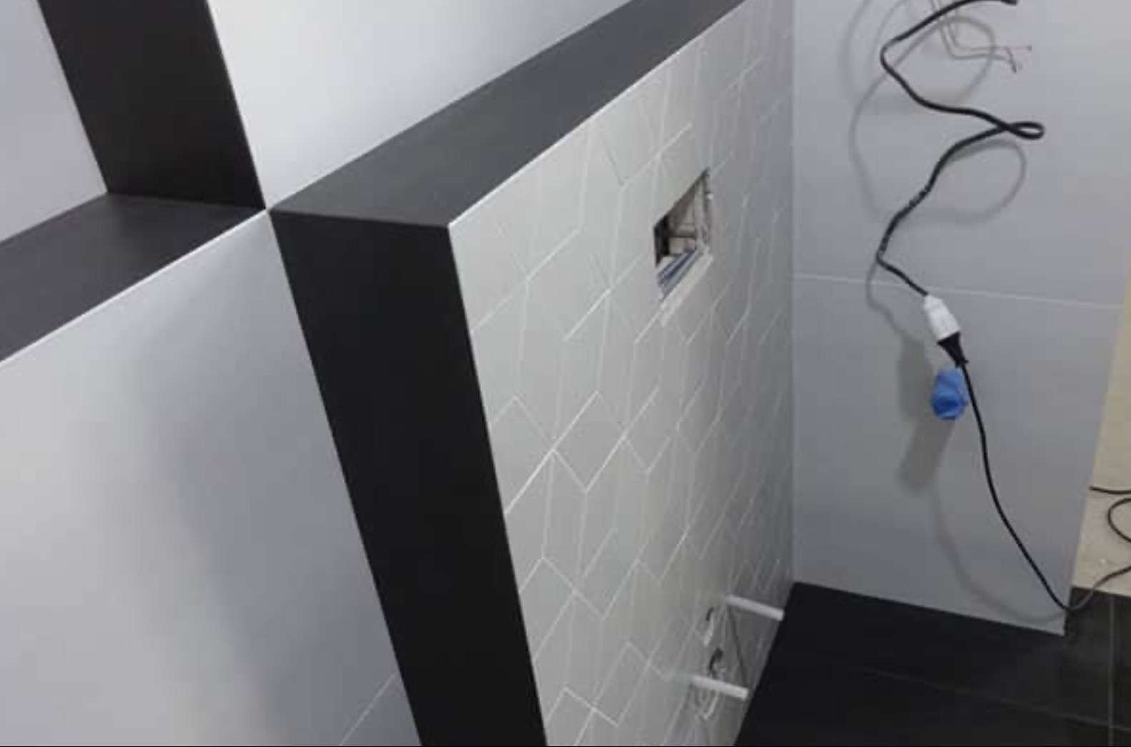 Ściana łazienki z geometrycznym wzorem płytek, czarnymi obramowaniami i wystającymi rurami instalacyjnymi. Widoczny fragment podłogi z czarnych płytek i przewód zasilający.