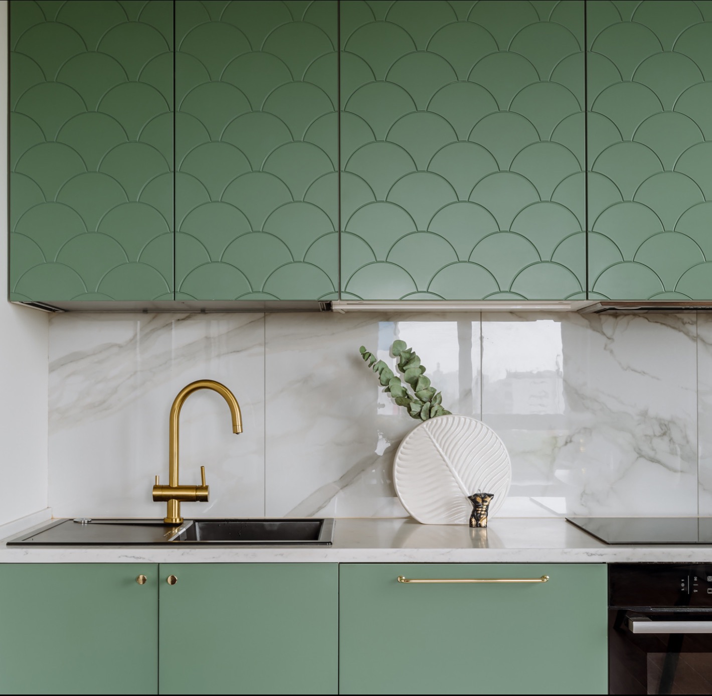 Stylowa kuchnia z zielonymi szafkami o wzorze rybiej łuski, złotą armaturą i blatem z marmuru. Nowoczesny design z dekoracyjną ceramiką i roślinami.