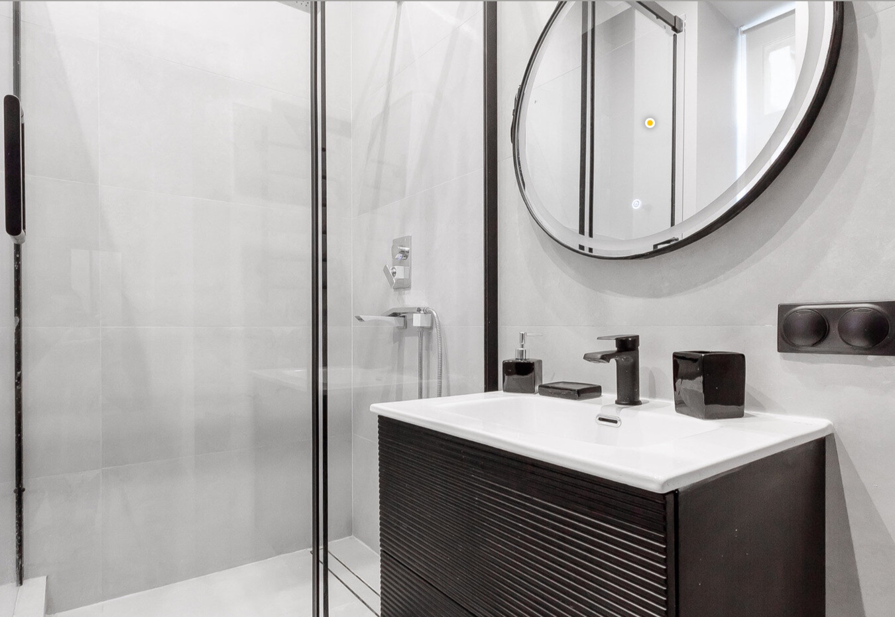Nowoczesna łazienka z kabiną prysznicową, okrągłym lustrem z podświetleniem i czarną armaturą. Minimalistyczny design, jasne płytki, czarne akcenty.