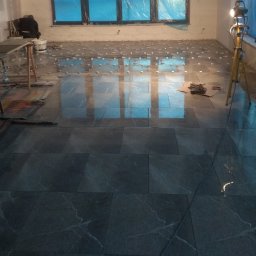 FHU Krzysztof Carukowski - Układanie ciemnych, marmurkowych płytek podłogowych w pomieszczeniu z oknami zabezpieczonymi folią, widoczny pracownik w tle i lampa budowlana na trójnogu.