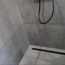 Nowoczesna kabina prysznicowa z szarymi płytkami imitującymi beton i czarnym odpływem liniowym w rogu.