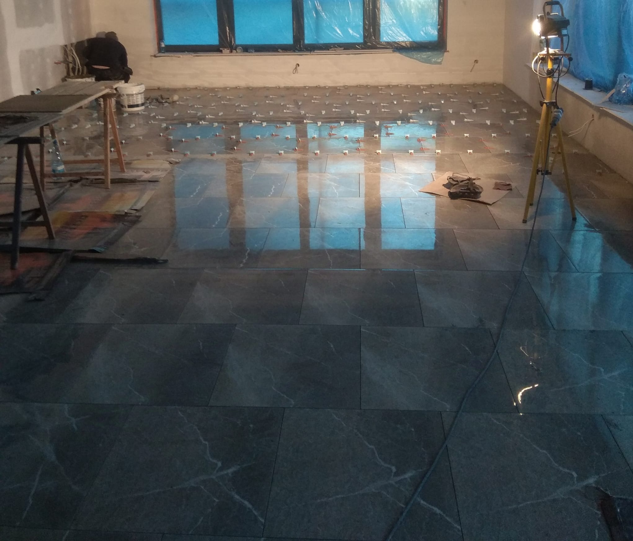 Układanie ciemnych, marmurkowych płytek podłogowych w pomieszczeniu z oknami zabezpieczonymi folią, widoczny pracownik w tle i lampa budowlana na trójnogu.