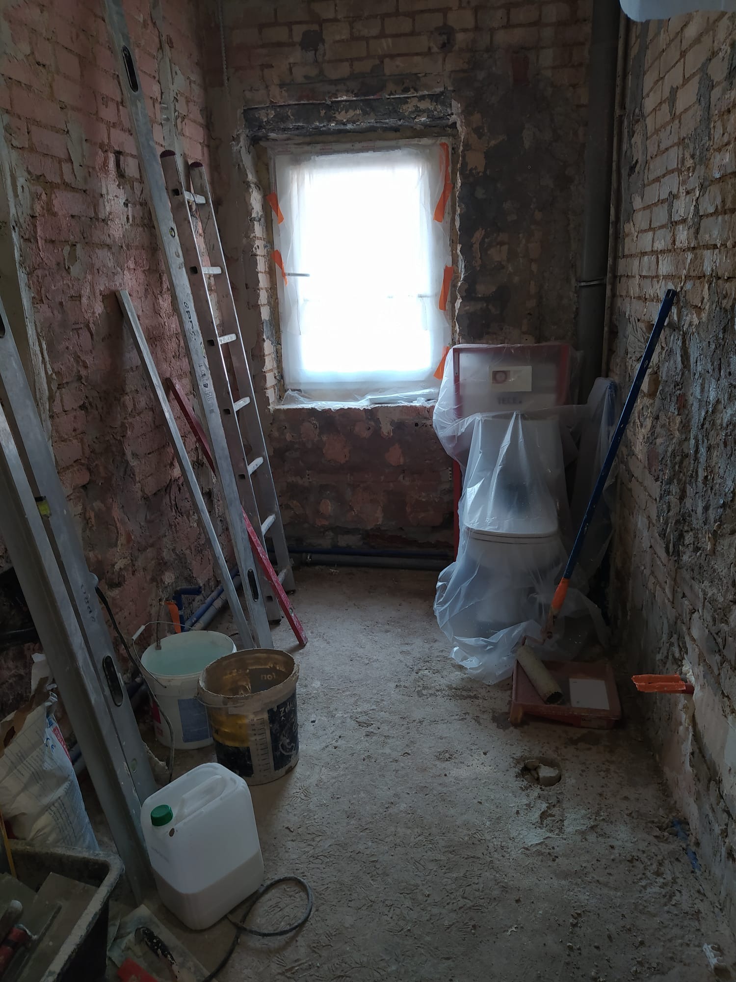 Wnętrze łazienki w trakcie remontu: odsłonięta cegła, drabiny opierają się o ścianę, toaleta zabezpieczona folią, narzędzia i pojemniki na podłodze, okno zasłonięte materiałem.