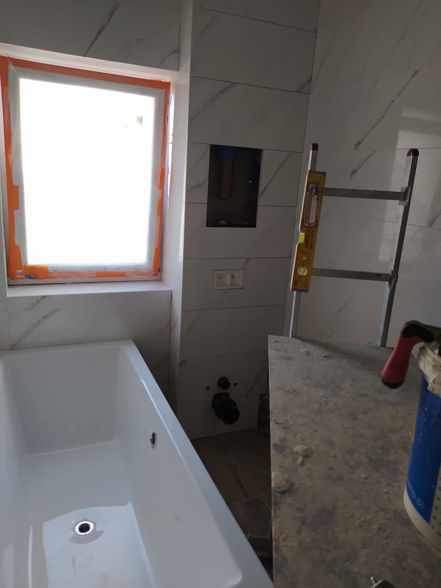 Biała wanna w trakcie montażu w łazience z marmurowymi płytkami, okno zabezpieczone taśmą malarską, widoczna poziomica i drabina.