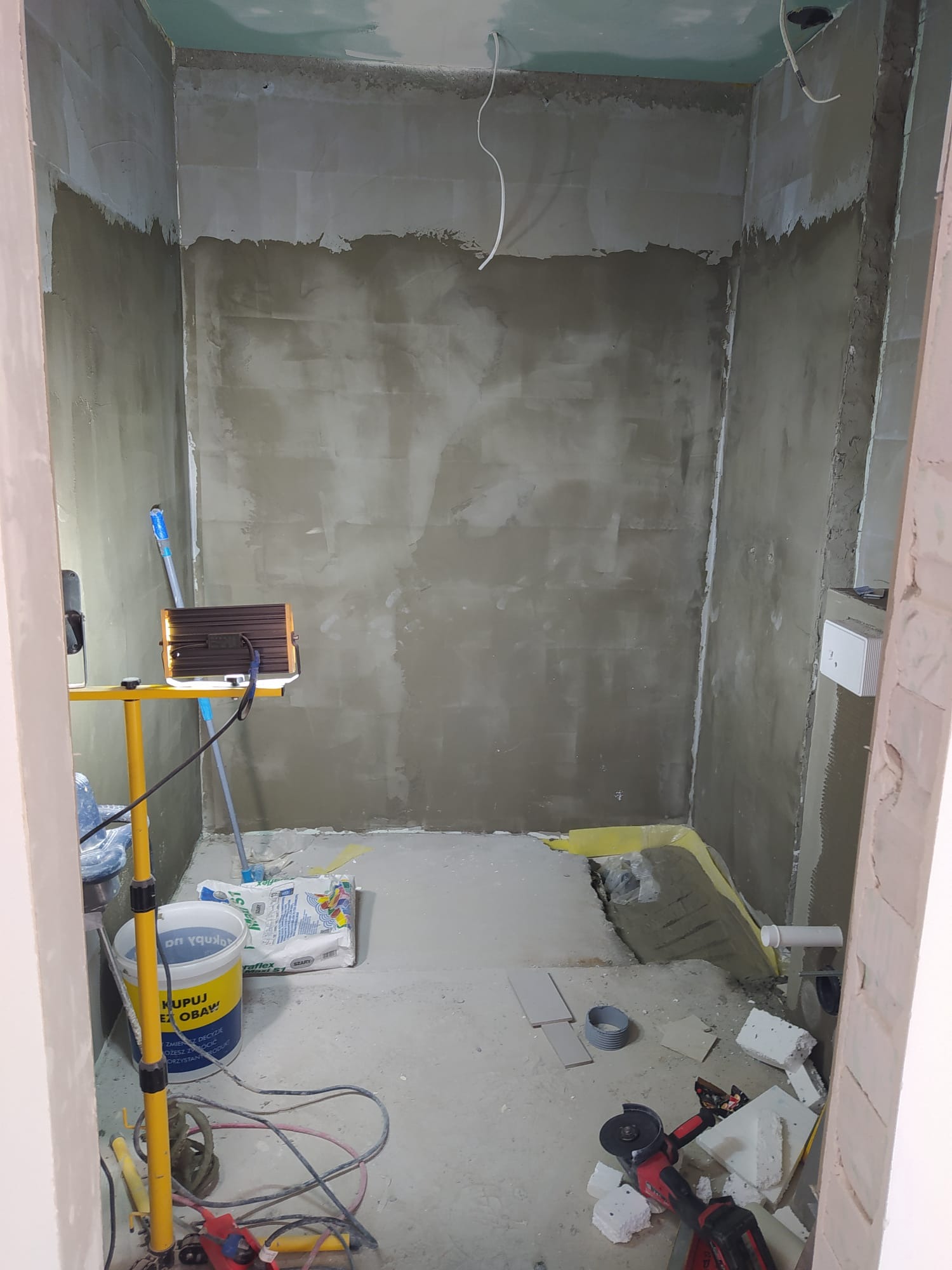 Łazienka w trakcie remontu: surowe ściany z widocznymi śladami tynku, narzędzia budowlane, oświetlenie robocze na żółtym statywie, przygotowane płytki na podłodze i wystające rury kanalizacyjne.