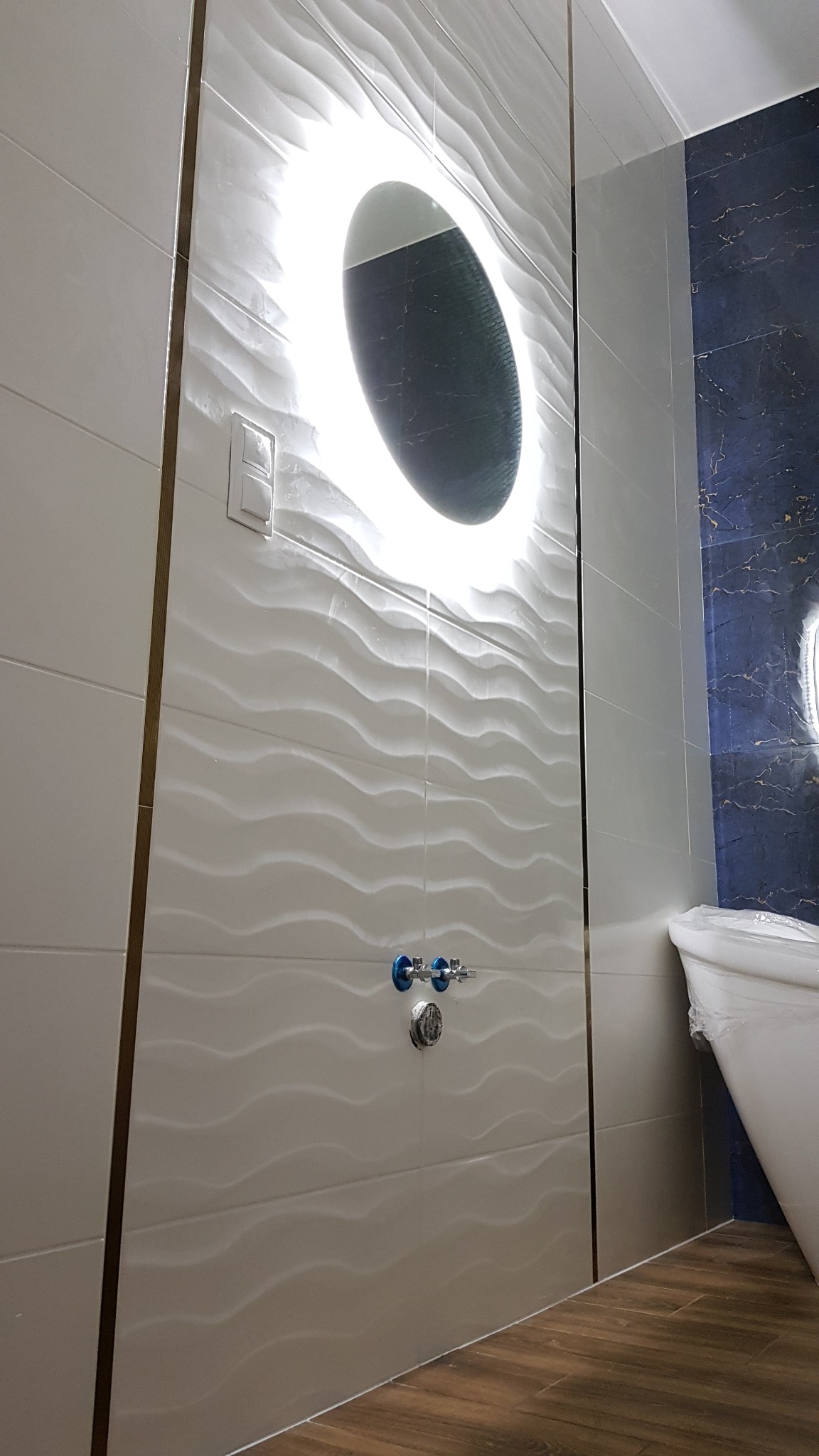 Białe, faliste płytki w łazience z okrągłym lustrem podświetlanym LED, widoczne zawory doprowadzające wodę, fragment toalety zabezpieczonej folią i ciemnoniebieska ściana w tle.