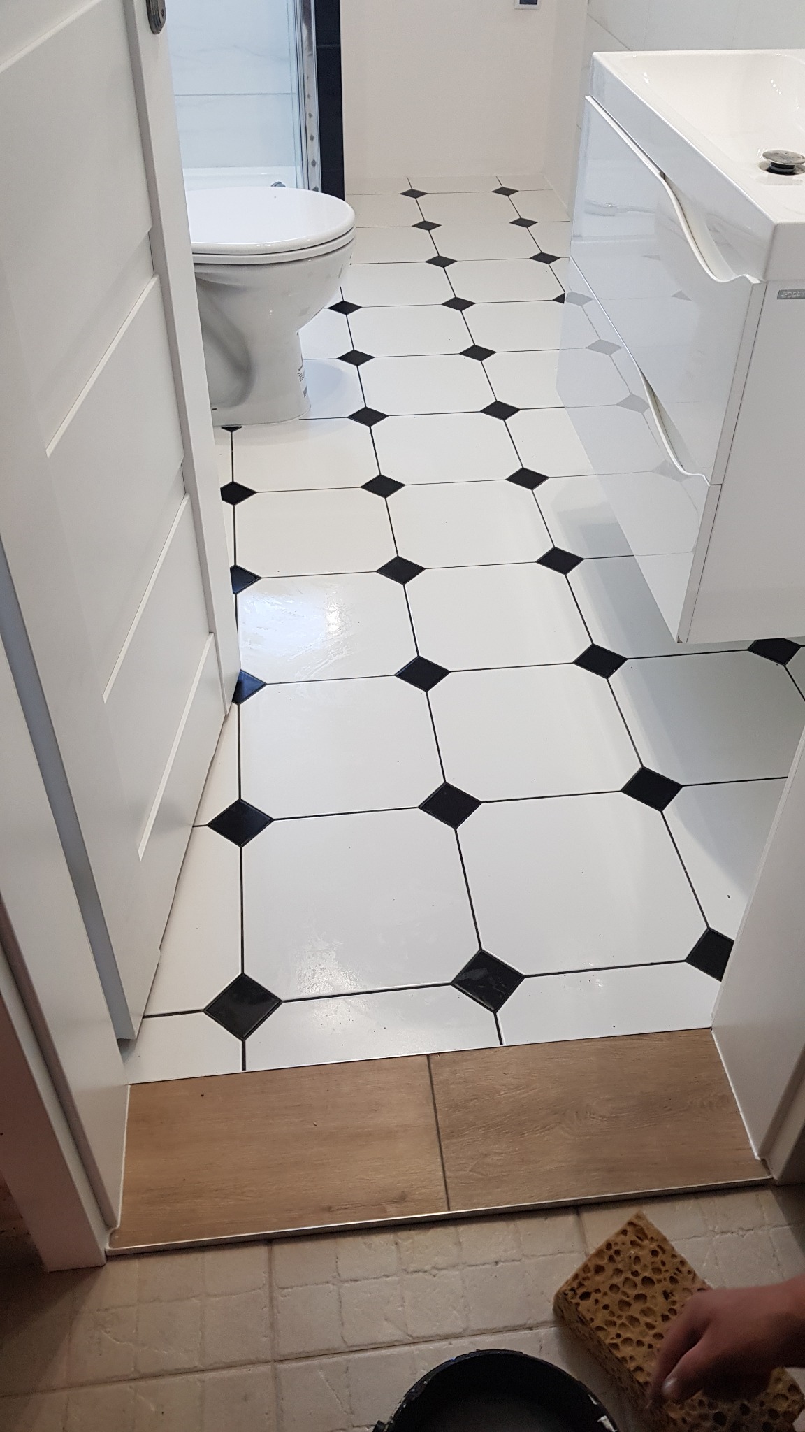 Biało-czarna podłoga w łazience z nową armaturą, widok z uchylonych drzwi, z widoczną gąbką i wiadrem na podłodze.