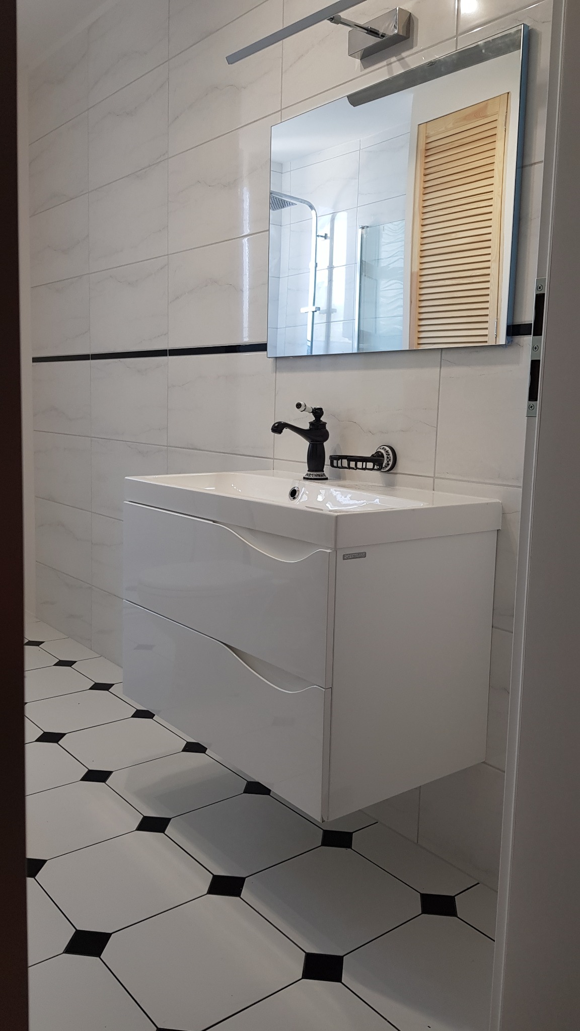 Biała szafka pod umywalkę z czarną baterią w łazience z biało-czarną podłogą w karo, widok na lustro z odbiciem kabiny prysznicowej i drewnianych żaluzji.
