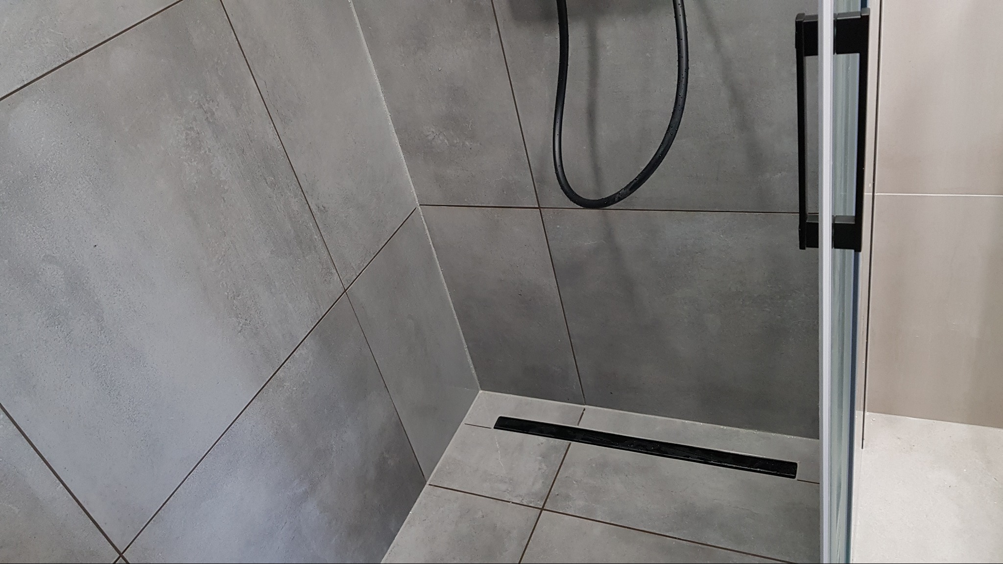 Nowoczesna kabina prysznicowa z szarymi płytkami imitującymi beton i czarnym odpływem liniowym w rogu.
