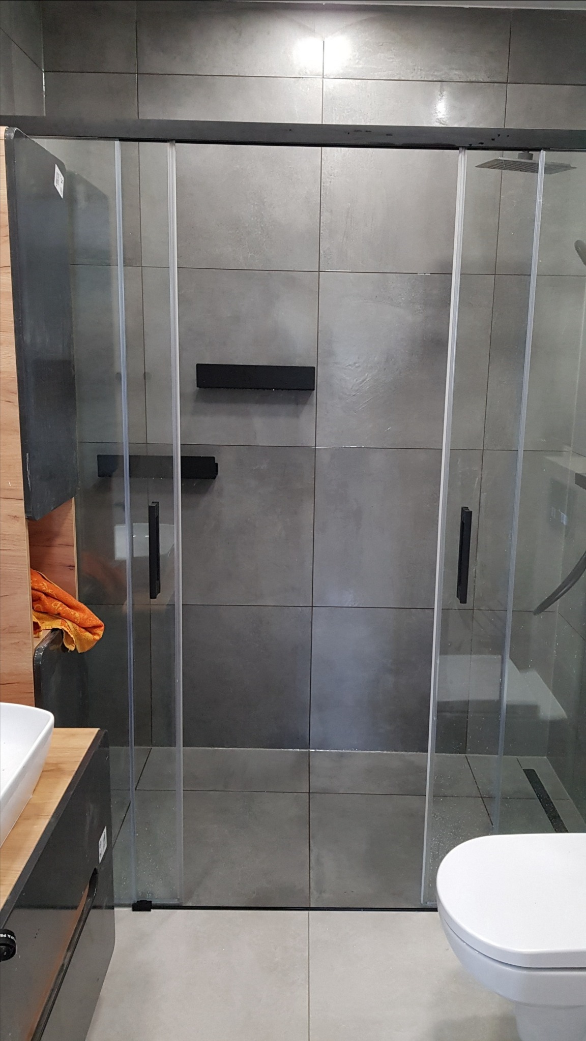 Szklana kabina prysznicowa z przesuwnymi drzwiami, szare płytki na ścianie, czarne akcenty armatury, widoczna toaleta i umywalka z szafką.