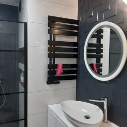 Usługi remontowo-budowlane Grzegorz Iskra - Nowoczesna łazienka w Zielonej Górze z czarno-białymi płytkami, okrągłym lustrem z podświetleniem LED, umywalką nablatową i czarnym grzejnikiem drabinkowym z różowym ręcznikiem.