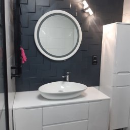 Usługi remontowo-budowlane Grzegorz Iskra - Nowoczesna łazienka z umywalką nablatową, okrągłym lustrem z podświetleniem LED, ciemną ścianą z geometrycznym wzorem płytek i białymi szafkami.