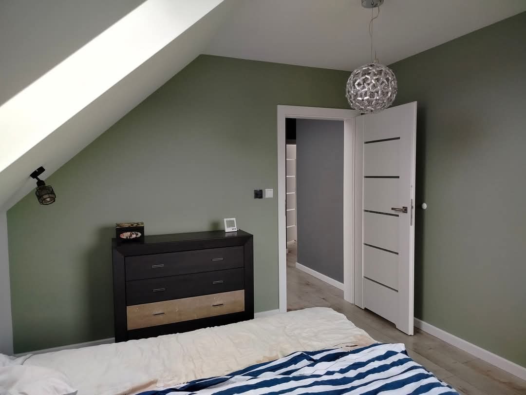 Pokój na poddaszu z oliwkowymi ścianami, czarną komodą z drewnianą szufladą, białą lampą kulą i otwartymi białymi drzwiami z czarnymi paskami.