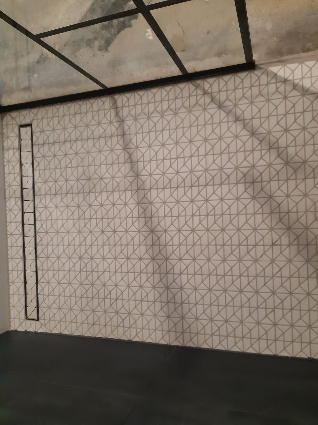 Ściana wykończona płytkami z geometrycznym wzorem trójkątów w odcieniach szarości, ciemna podłoga i sufit z widoczną instalacją.