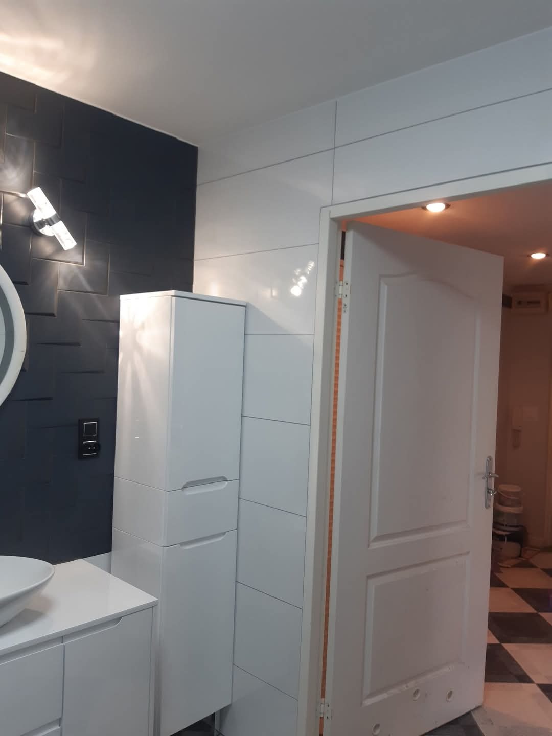 Wykończona łazienka z białą armaturą, czarną ścianą z dekoracyjną fakturą i otwartymi drzwiami, przez które widać fragment szachownicy na podłodze.