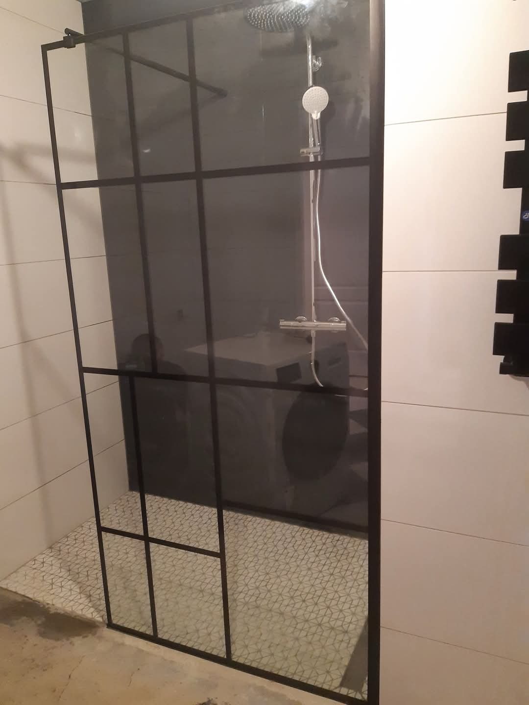 Kabina prysznicowa z czarnymi profilami, widoczna bateria i deszczownica, w tle pralka, płytki podłogowe z geometrycznym wzorem.