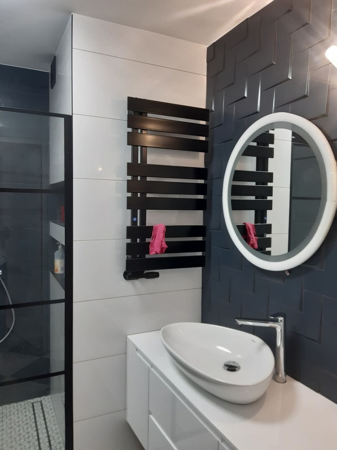 Nowoczesna łazienka w Zielonej Górze z czarno-białymi płytkami, okrągłym lustrem z podświetleniem LED, umywalką nablatową i czarnym grzejnikiem drabinkowym z różowym ręcznikiem.