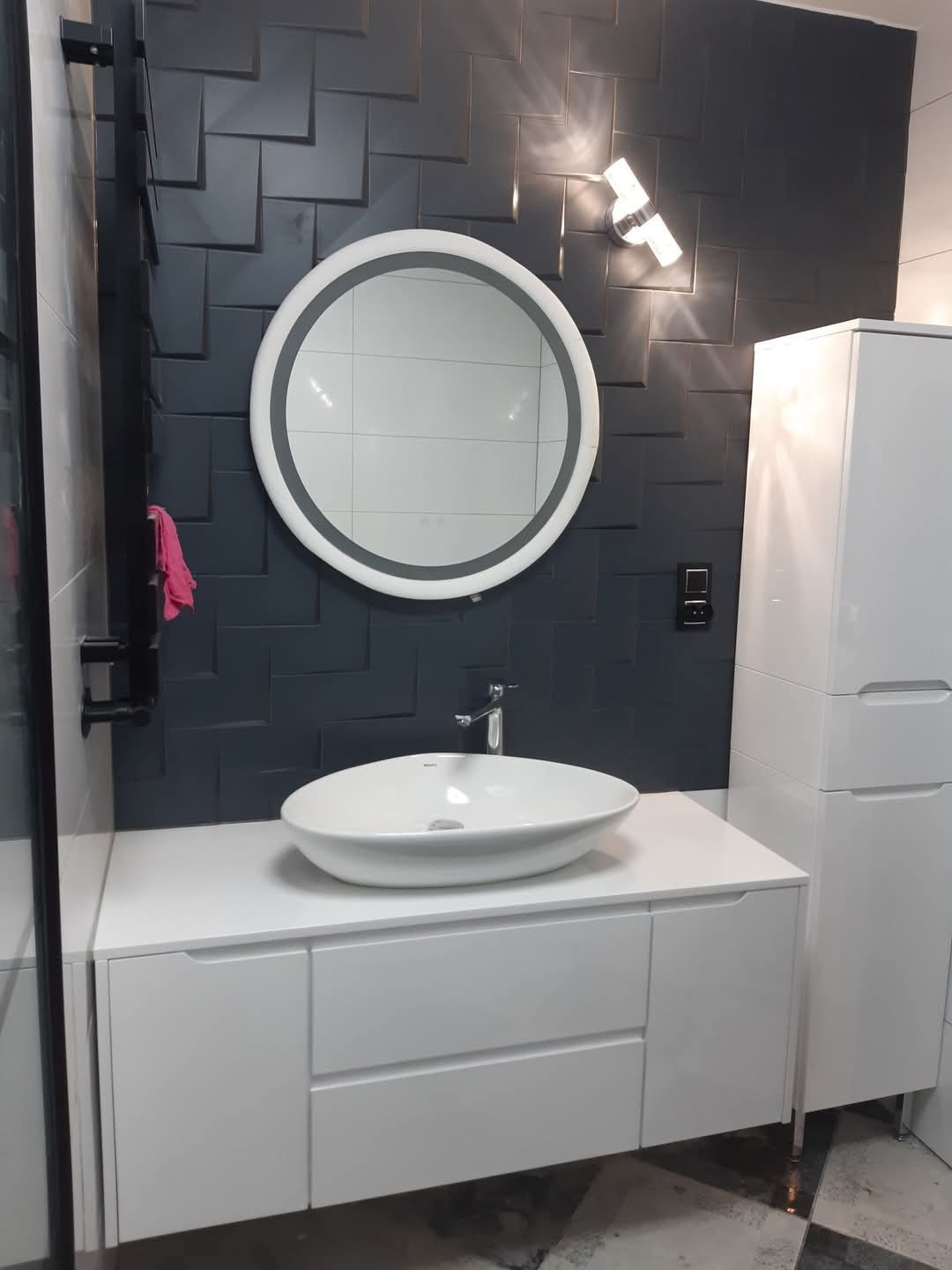 Nowoczesna łazienka z umywalką nablatową, okrągłym lustrem z podświetleniem LED, ciemną ścianą z geometrycznym wzorem płytek i białymi szafkami.