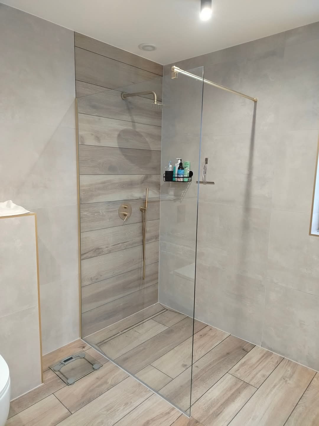 Nowoczesna kabina prysznicowa walk-in z szklaną ścianką, wykończona płytkami imitującymi drewno w odcieniach szarości i beżu, z złotymi elementami armatury i minimalistyczną półką na kosmetyki...